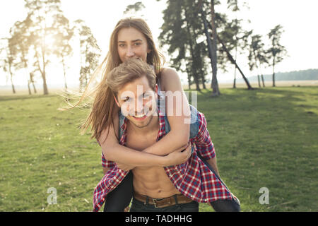Portrait of happy young man carrying girlfriend piggyback on meadow Stock Photo