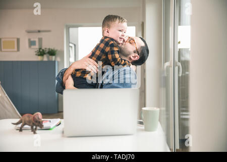 Father sitting at table in home office hugging son Stock Photo