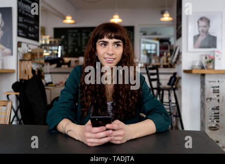 Smiling young woman with cell phone in a cafe looking around Stock Photo