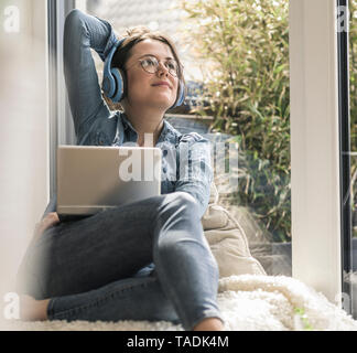 Woman with headphones and laptop sitting at the window at home Stock Photo