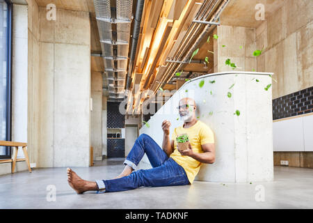 Mature businessman sitting on the floor in modern office surrounded by floating lettuce leaves Stock Photo