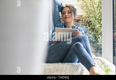 Woman with tablet sitting at the window at home with closed eyes Stock Photo