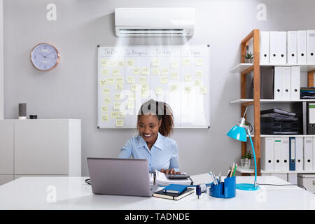 Smiling Businesswoman Enjoying The Cooling Of Air Conditioner Using Laptop At Workplace Stock Photo