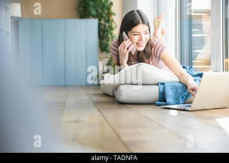 Young woman lying at the window at home using cell phone and laptop Stock Photo