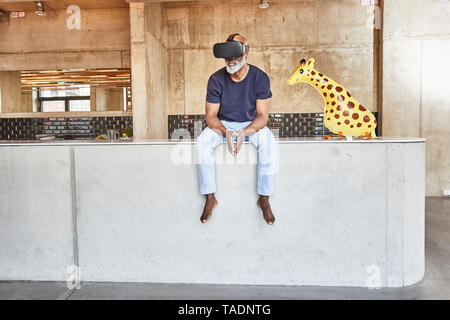 Mature businessman in modern office sitting next to giraffe figurine wearing VR glasses Stock Photo