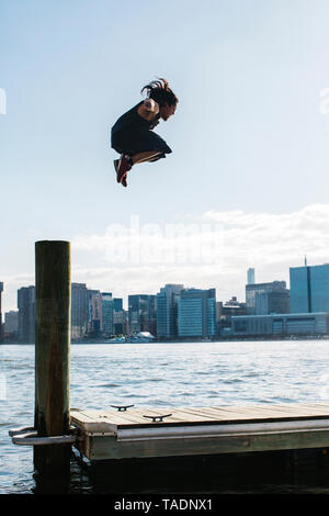 USA, New York, Brooklyn, young man doing Parkour jump from wooden pole in front of Manhattan skyline Stock Photo