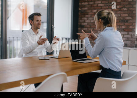Businessman and woman sitting at desk, working on laptop, pointing with pistol fingers at each other Stock Photo