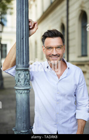 Portrait of smiling businessman leaning against lamp post in the city Stock Photo