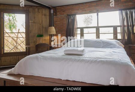 White Bed in Bedroom of Interior Design Room in Country Loft Style. Interior Design Room include wood chair and table and lamp and windows on brick wa Stock Photo