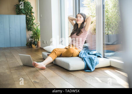 Relaxed young woman sitting at the window at home with laptop Stock Photo