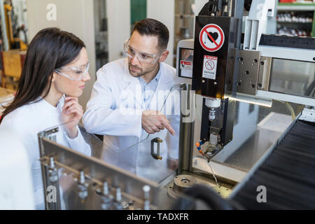 Colleagues wearing lab coats and safety goggles at machine in modern factory Stock Photo