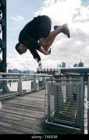 USA, New York, Brooklyn, young man doing Parkour handstand on railing of pier in front of Manhattan skyline