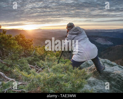Hiker and photo enthusiast stay with tripod on cliff. Peak with woman taking photos in morning sunrise Stock Photo