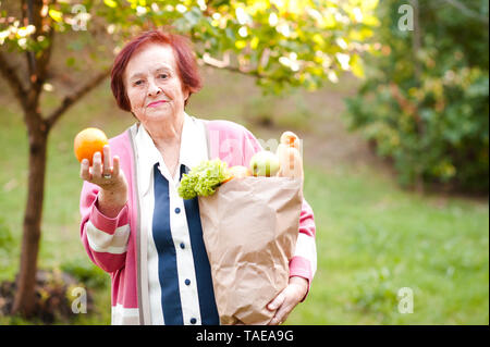 Smiling senior woman 70-80 year old holding paper bag with food outdoors. Looking at camera. Stock Photo
