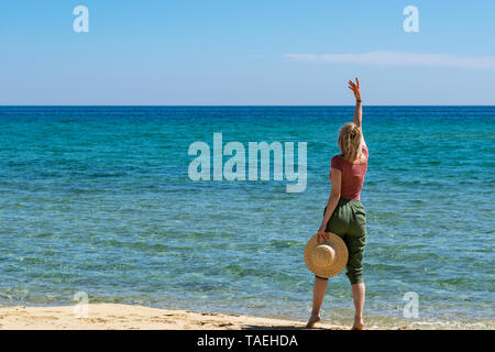 Rear view of young woman with casual outfit and straw hat in hand standing with one arm raised at beach against clear sky and blue sea Stock Photo