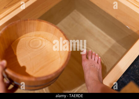 Cypress bathtub wooden traditional Japanese bucket by tub in onsen hotel bathroom interior in Japan with feet stepping into water Stock Photo