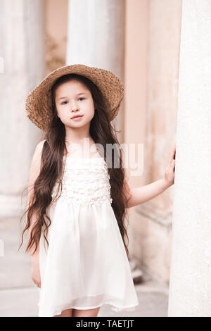 Cute kid girl 4-5 year old wearing straw hat and stylish white dress outdoors. Walking in ancient part of city. Looking at camera. Stock Photo