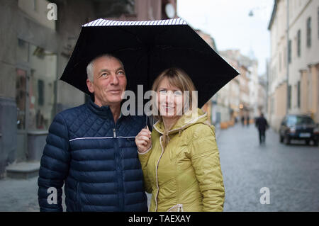 Middle-aged woman and her elderly husband spending time together outdoors standing under their umbrella on paved street. Couple with age difference Stock Photo