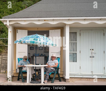 Older retired couple sitting on beach chairs recliners outside their beach hut in Bournemouth near Middle Chine and Alum Chine under umbrella Stock Photo
