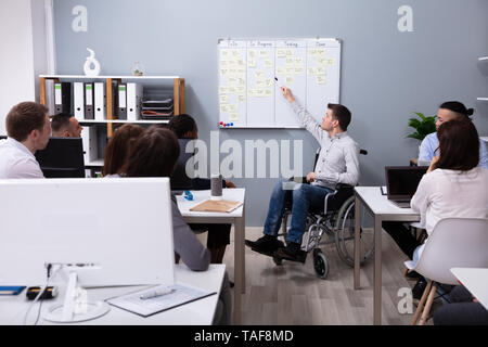 Disabled Young Businessman On Wheelchair Giving Presentation To His Colleague In Office Stock Photo