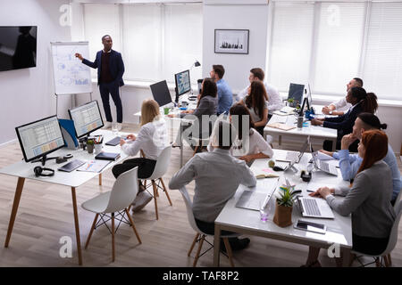 African Businessman Giving Presentation To His Colleagues Sitting On Table With Computers In Office Stock Photo