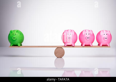 Green And Pink Piggy Bank Balancing On Wooden Seesaw Over Reflective Desk Stock Photo