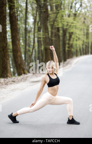 A young, sympathetic blonde woman is actively spending time outdoors, doing a warm up before jogging. Stock Photo