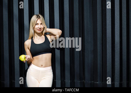 A young, sympathetic blonde woman is actively spending time outdoors, holding a green apple. The girl is dressed in sportswear. Time for a healthy sna Stock Photo