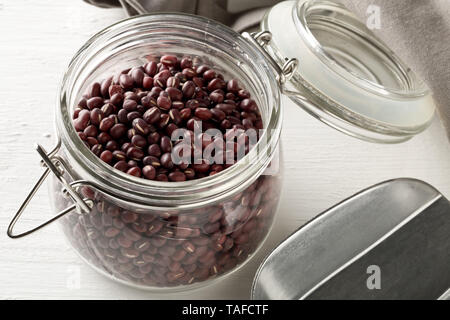 Raw, uncooked, dried adzuki (red mung) beans in glass storing jar with metal spoon on white wood table background with grey kitchen towel Stock Photo