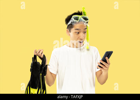 Korean young man's half-length portrait on yellow studio background. Male model in white shirt and swimglasses. Holding flippers. Concept of human emo Stock Photo