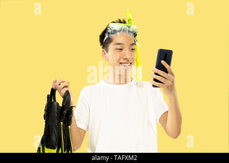 Korean young man's half-length portrait on yellow studio background. Male model in white shirt and swimglasses. Holding flippers. Concept of human emo Stock Photo