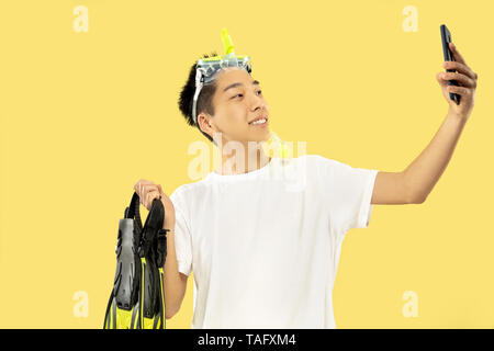 Korean young man's half-length portrait on yellow studio background. Male model in white shirt and swimglasses. Holding flippers. Concept of human emo Stock Photo