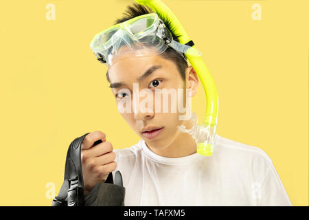 Korean young man's half-length portrait on yellow studio background. Male model in white shirt and swimglasses. Holding flippers. Concept of human emo Stock Photo