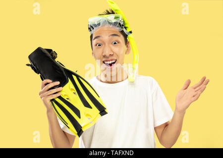 Korean young man's half-length portrait on yellow studio background. Male model in white shirt and swimglasses. Holding flippers. Concept of human emo Stock Photo