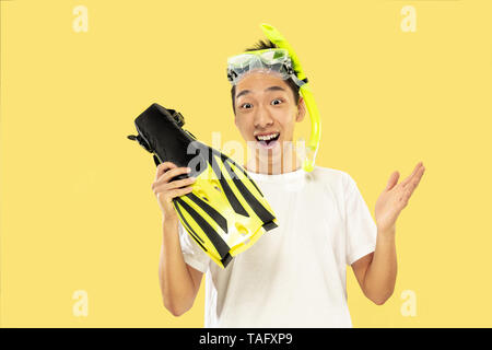 Korean young man's half-length portrait on yellow studio background. Male model in white shirt and swimglasses. Holding flippers. Concept of human emo Stock Photo