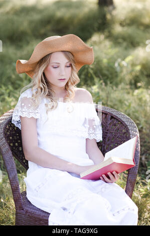 Beautiful blonde girl 15-16 year old reading book sitting in chair outdoors. Stock Photo