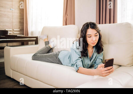 Photo of relaxed caucasian woman watches online story on mobile phone, reads news with amazing information, dressed in casual home clothes, lies on so Stock Photo