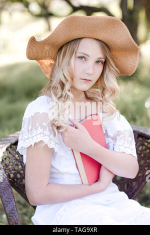 Beautiful blonde girl 14-16 year old holding book sitting in chair in park. Looking at camera. Stock Photo