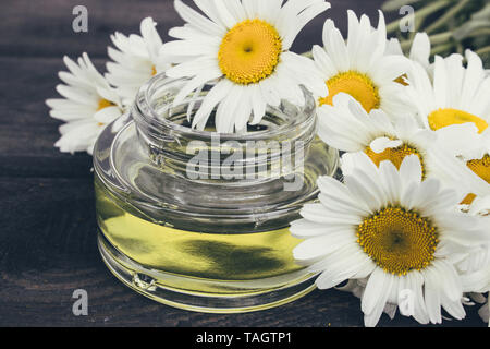A can of light essential oil from chamomiles stands on dark boards near chamomile flowers. Stock Photo
