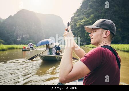 Tourist with mobile phone on boat. Young man photographing of river against karst formation near Tam Coc in Ninh Binh province, Vietnam. Stock Photo