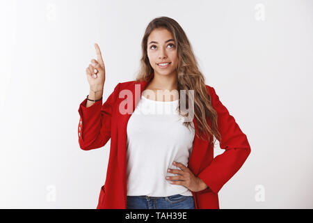 Cute dreamy young 25s millennial curly-haired female wearing red jacket looking pointing up smiling curiously amused, high-hopes cheering up self Stock Photo