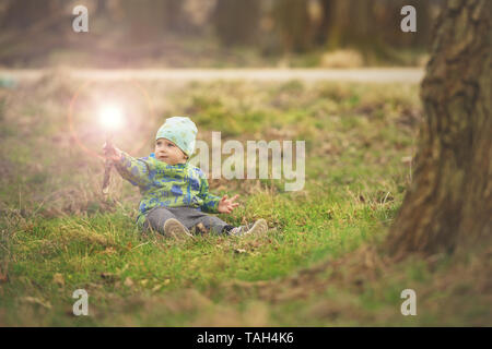 Small boy is sitting on grass and handling magic wand in spring park near big tree Stock Photo