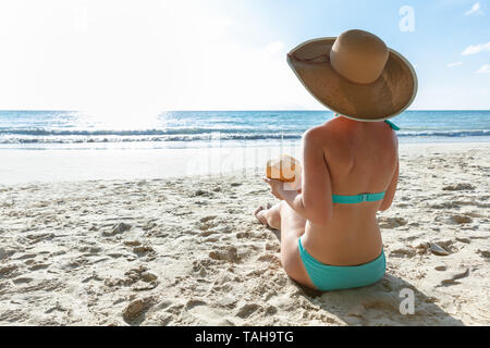 Rear View Of A Woman In Bikini Hat Sitting On Sand Holding Coconut At Beach Stock Photo