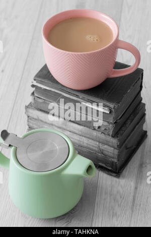 Cup of tea in a pink cup with pastel green teapot and a stack of old books with selective colour on a grey wood background Stock Photo