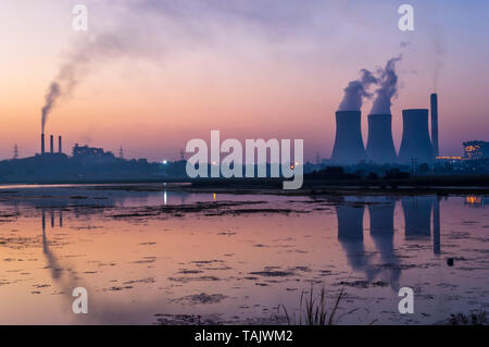 Coal powered thermal power plant emitting smoke and steam from chimney and cooling tower. Reflection of cooling tower and chimney on lake. Stock Photo