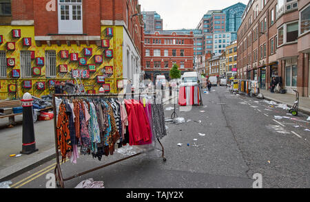 LONDON SPITALFIELDS BRICK LANE AREA STREET AT THE END OF A MARKET DAY WITH ART ON A LARGE BRICK WALL AND RACKS OF CLOTHES IN THE ROAD Stock Photo