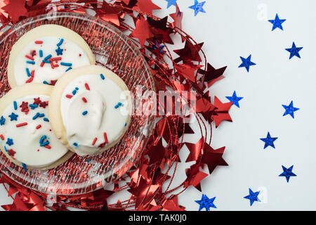 bright shiny red star garland under a glass plate with sugar cookies covered with white frosting and sprinkles and blue glitter stars on a white table Stock Photo