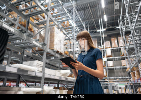 Wholesale warehouse. Beautiful young woman worker of store in shopping center. Girl looking for goods with a tablet is checking inventory levels in a Stock Photo