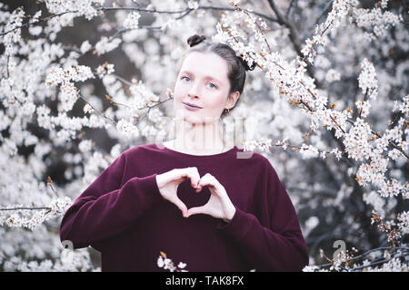 Beautiful smiling girl 20-24 year old making heart shape with hands outdoors over blooming tree. Looking at camera. Spring time. Stock Photo