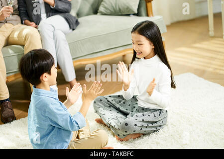 two cute little asian kids brother and sister sitting on carpet playing game with parent sitting on sofa in the background Stock Photo